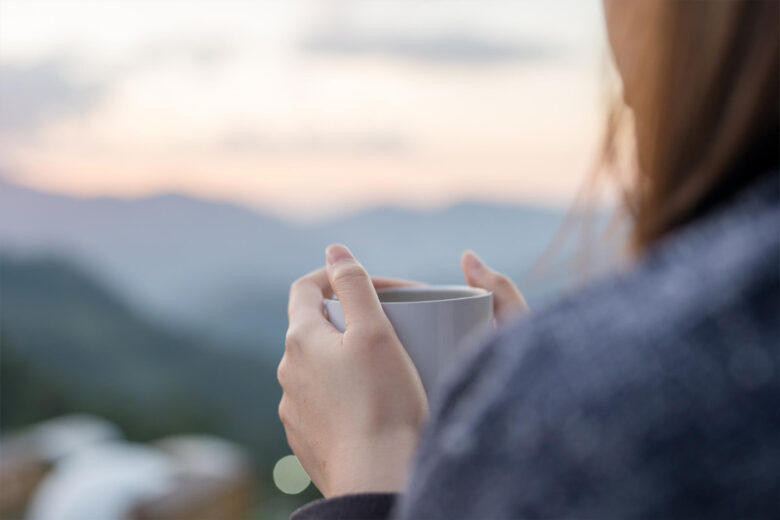 山の景色を見ながら暖かい飲み物を飲んでくつろぐ女性の後ろ姿